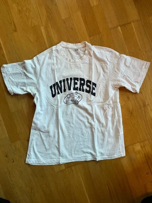 Vit t-shirt med tryck 'UNIVERSE' - Vit t-shirt med svart text 'UNIVERSE' och ett tryck av en planet på bröstet. Klassisk passform med rund hals och korta ärmar. Perfekt för dig som gillar enkel men cool stil.