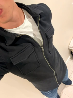 Svart overshirt med dragkedja - Säljer en svart overshirt med dragkedja framtill och två stora bröstfickor med lock. Overshirten har klassisk krage och är gjord i ett mjukt, tjockare tyg som ger en clean och stilren look. Perfekt att slänga över en t-shirt för en avslappnad vibe.