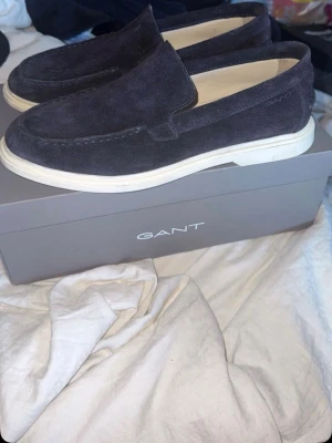 Mörkblå mockaloafers från GANT - Stilrena loafers i mörkblå mocka från GANT med vit sula. Skorna har en klassisk slip-on design och är tillverkade i mjukt mockamaterial som ger en lyxig känsla. Perfekta för dig som gillar en clean och avslappnad look.