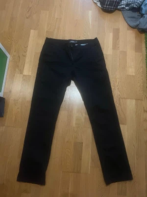 Svarta chinos med rak passform - Säljer ett par svarta chinos med rak passform och klassisk design. Byxorna har framfickor, bakfickor med knapp och stängs med dragkedja och knapp i midjan. Perfekta för en clean och stilren look.