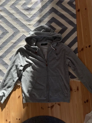 Grå hoodie med dragkedja från Morris - Snygg grå hoodie från Morris med dragkedja framtill, två fickor och vit dragsko i huvan. Klassisk passform och diskret logga på bröstet. Perfekt för en avslappnad och stilren look. Pris kan diskuteras.