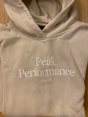 Beige hoodie från Peak Performance - Snygg beige hoodie från Peak Performance med broderad vit logga och texten 'Est. 1986' på bröstet. Klassisk modell med huva och magficka. Perfekt för en avslappnad och trendig stil. Mjuk bomullsblandning som känns skön mot huden.