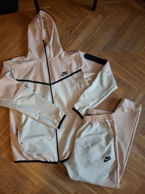 Nike beige loungewear set med huva - Nike loungewear set i beige med svarta detaljer. Setet består av en zip-hoodie med huva och svarta linjer samt matchande byxor med resår i midjan. Båda plaggen har Nike-logga framtill. Perfekt för chill dagar hemma.