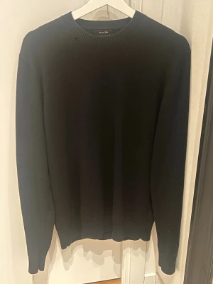 Svart stickad tröja Massimo Dutti - Klassisk svart stickad tröja från Massimo Dutti i en blandning av ull och bomull. Tröjan har rund halsringning, långa ärmar och ribbade muddar vid ärmslut och nederkant. Perfekt för en stilren och enkel look. Hål vid kragen men annars i mycket bra skick!
