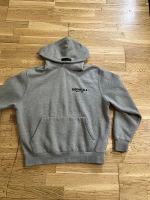 Grå Essentials hoodie med tryck - Snygg grå hoodie från Essentials med svart tryck på bröstet. Klassisk modell med huva och stor magficka framtill. Mjuk bomullsblandning och ribbade muddar vid ärmslut och nederkant. Perfekt för en avslappnad streetwear-look.