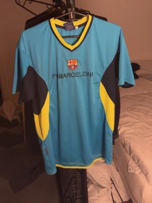 Retro barca träningskit - Snygg blå fotbollströja från FC Barcelona med klubbmärke och text på bröstet. Tröjan har gula och mörkblå detaljer samt gul kant vid halsen. Perfekt för dig som vill representera ditt favoritlag på planen eller till vardags.