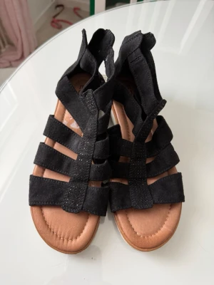 Svarta gladiatorsandaler i mocka - Stilrena svarta gladiatorsandaler med flera breda remmar i mocka och små diskreta nitar på mittenremmen. Sandalerna har en mjuk, ljusbrun innersula och vit yttersula. Perfekta för en trendig och bekväm look under varma dagar.