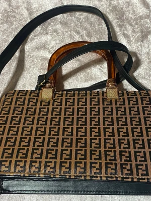 Dam  brun handväska / Axelväska med FF-mönster - Snygg handväska i brunt och svart med klassiskt FF-mönster över hela väskan. Väskan har bärhandtag i brun plast, svarta detaljer . Perfekt för dig som gillar statement-accessoarer.