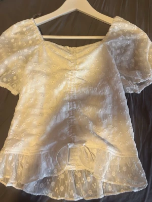 Vit blus med volang från Lindex - Söt vit blus från Lindex med korta puffärmar, broderade blommor och volangkant nedtill. Toppen har snörning framtill och är tillverkad i ett lätt, luftigt material som passar perfekt till sommaren.