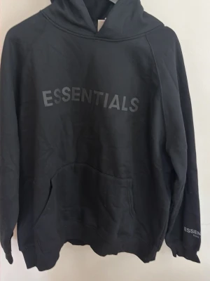 Svart Essentials hoodie med huva - Säljer en svart hoodie från Fear of God Essentials med stor logga på bröstet och mindre logga på ärmen. Hoodien har en rymlig huva och en klassisk känguruficka framtill. Tillverkad i mjukt bomullsmaterial som är perfekt för en chill och avslappnad stil.