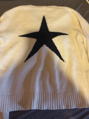 Vit stickad tröja med svart stjärna - Vit stickad tröja med en stor svart stjärna på ryggen. Tröjan har v-ringning, långa ärmar och breda ribbade muddar vid ärmslut och nederkant. Perfekt för dig som gillar enkel men cool design.