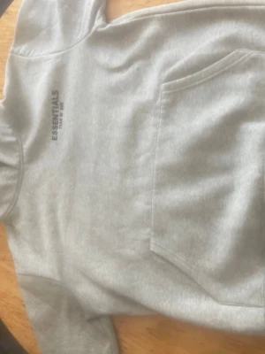 Grå Essentials Fear of God hoodie - Snygg grå hoodie från Essentials Fear of God med svart logga på bröstet. Klassisk känguruficka framtill och huva. Perfekt för en avslappnad streetwear-look. Mjuk bomullsblandning som känns skön mot huden.