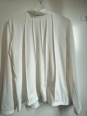 Vit polotopp med veckade detaljer - Vit långärmad polotopp med snygga veckade detaljer framtill. Topp i mjukt material med hög hals, perfekt för lager-på-lager. Enkel och stilren design som passar till det mesta. (Finns också i svart) 