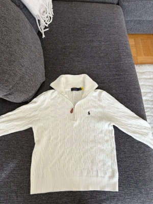 Vit stickad Ralph Lauren quater zip - Kabelstickad vit tröja från Polo Ralph Lauren med halv dragkedja och hög krage. Tröjan har det klassiska broderade logotypen på bröstet och ribbade muddar vid ärmslut och nederkant. Perfekt för en stilren och avslappnad look. Köparen betalar frakten 