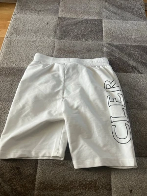 Vita Moncler shorts med tryck - Snygga vita shorts från Moncler med svart tryck längs benen som bildar varumärkets namn. Shortsen har elastisk midja med snörning och två sidofickor. Tillverkade i mjuk bomull, perfekta för sommaren.