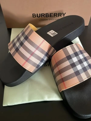 Burberry sandaler med rutigt band - Snygga sandaler från Burberry med klassiskt beige, svart och rött rutmönster på bandet. Sulan är svart och har en enkel, stilren design. Perfekta för sommaren och ger en lyxig touch till din outfit.