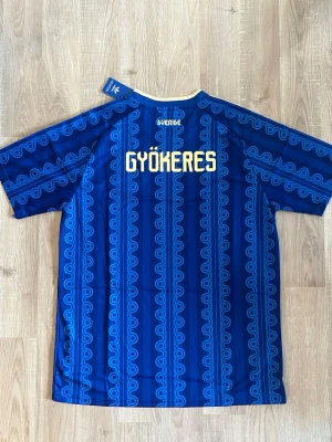 Sverige Gyökeres VM-2026 Fotbollströja - Säljer Sveriges Bortatröja med Sveriges landslagsemblem och namnet Gyökeres på ryggen. Tröjan har gula detaljer, klassiska Adidas-ränder på axlarna och ett unikt mönster över hela tyget. Tillverkad i lätt och ventilerande material. HELT NY