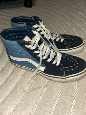 Blåa Vans High sneakers - Säljer ett par klassiska Vans High sneakers i blått och svart med vit sula och vit sidostripes. Skorna har högt skaft, vita snören och kontrastsömmar. Ovandelen är i canvas och mocka för en retro och streetig vibe. Storlek: 39