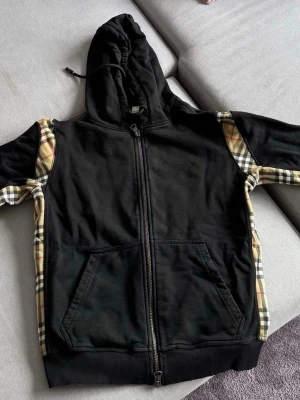 Svart hoodie med rutiga detaljer burberry - Säljer en svart hoodie med dragkedja och huva. Ärmar och sidor har klassiska beige, röda och svarta rutiga paneler som ger en snygg kontrast. Två fickor framtill och ribbade muddar. Perfekt för dig som gillar streetwear och vill sticka ut.
