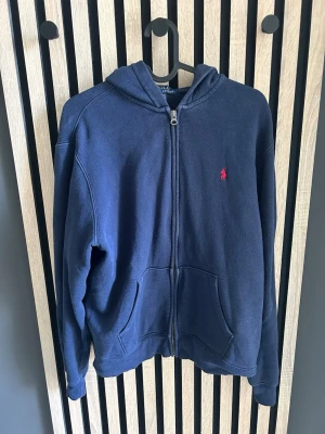 Marinblå hoodie från Polo Ralph Lauren -  Säljer en klassisk marinblå hoodie med dragkedja från Polo Ralph Lauren. Hoodien har två fickor framtill och den ikoniska röda broderade loggan på bröstet. Tillverkad i mjukt bomullsmaterial och har en avslappnad passform med huva. Kontakta mig vid frågor 😃