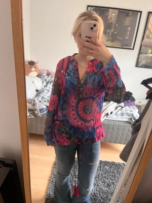 Desigual blommig blus - Säljer en färgstark blus med blommigt och mandalainspirerat mönster i rosa, blått och lila. Blusen har V-ringning med knappar och knytband i midjan. Långärmad modell i ett tunt, lätt material som känns luftigt och skönt.