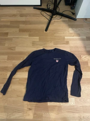 Mörkblå långärmad tröja från GANT - Mörkblå långärmad tröja från GANT med rund halsringning och diskret logga samt sköld på bröstet. Tröjan har en smal passform och är tillverkad i ett mjukt, tunt material som passar perfekt till lager på lager.