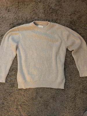 Grå stickad tröja  - Säljer en stilren beige stickad tröja från Maison Margiela. Tröjan har rund halsringning, ribbstickad struktur och långa ärmar. Perfekt för dig som gillar minimalistisk och tidlös design. Materialet känns mjukt och håller värmen bra.