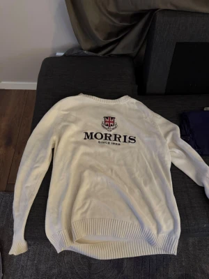 Vit tröja från Morris med tryck - Vit stickad tröja från Morris med broderad logga och brittisk flagga på bröstet samt texten 'Morris Since 1968'. Siffran 68 är broderad på ena ärmen. Tröjan har rund hals och ribbade muddar vid ärmslut och nederkant.