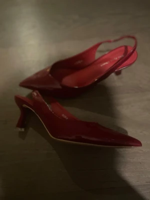 Röda lackpumps med öppen häl - Snygga röda pumps i lack med spetsig tå och öppen häl. Skorna har en smal rem runt hälen och en elegant, smal klack. Perfekta för dig som vill sticka ut med ett par glansiga och trendiga klackar. Materialet är glansigt och färgen är djupt röd.