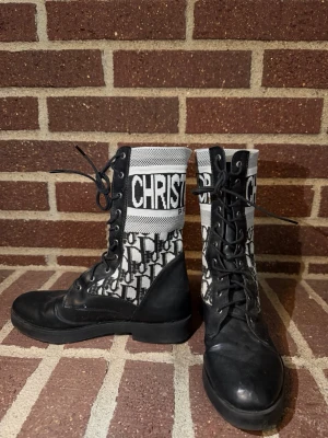 Svarta snörade stövletter Dior - Snygga svarta stövletter från Christian Dior med klassiskt monogrammönster i svart och vitt. Högt skaft med tydlig logga, snörning framtill och detaljer i både skinn och textil. Perfekta för dig som vill sticka ut med en lyxig och trendig look.