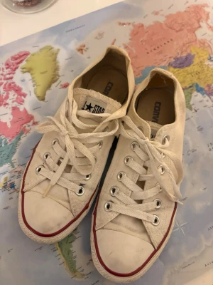 Vita Converse All Star sneakers - Klassiska vita Converse All Star sneakers i lågt skaft med snörning. Skorna har vit canvas, gummisula med röd och blå rand samt metallöljetter. Perfekta för en avslappnad och cool stil.