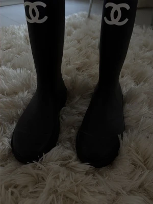 Chanel boots - Säljer ett par svarta höga gummistövlar från Chanel med den ikoniska vita CC-loggan framtill på skaftet. Stövlarna har en klassisk, stilren form och är perfekta för regniga dagar. Tillverkade i slitstarkt gummimaterial och har en rund tå.