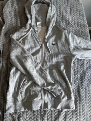 Grå och svart zip hoodie från Nike - Få båda två för 250! Snygg grå och svart zip hoodie från Nike med dragkedja framtill och klassisk logga broderad på bröstet. Hoodien har huva med dragsko och två fickor framtill. Perfekt för chill dagar eller när du vill ha en sportig look.