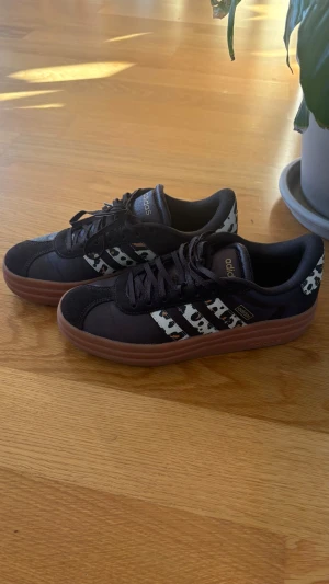 Adidas sneakers med leopard mönster  - Snygga Adidas sneakers i svart mocka med klassiska tre ränder i vitt och svart djurmönster. Skorna har brun gummisula och låg profil. Perfekta för dig som vill sticka ut med en unik detalj på fötterna.