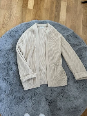Beige ribbstickad kofta från Arket - Snygg beige kofta från mango i ribbstickat material. Koftan har öppen front, långa ärmar och två fickor framtill. Perfekt för lager-på-lager och enkel att matcha med olika outfits.