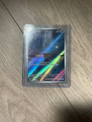 Pokémonkort Cleffa holografisk - Säljer ett holografiskt Pokémonkort med Cleffa-motiv. Kortet har en skimrande regnbågseffekt och ligger i ett genomskinligt plastfodral. Texten på kortet är på engelska och det har attacken Grasping Draw.