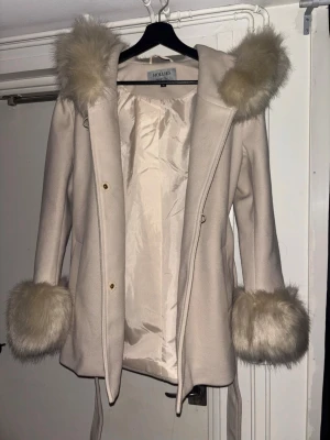 Beige kappa med fuskpäls från Holais - Stilren beige kappa från Hollies med luva och detaljer i ljus fuskpäls på både huva och ärmslut. Kappan har knäppning framtill, avtagbart midjebälte och slät insida. Pälsen är avtagbar runt ärmarna. Kappan är aldrig använd.