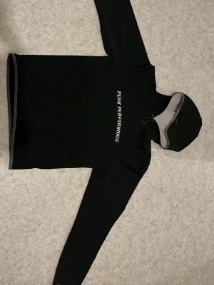 Svart hoodie Peak Performance - Svart hoodie från Peak Performance med vit logga på bröstet och justerbar huva. Tröjan har långa ärmar och gråa detaljer vid ärmslut och nederkant. Materialet är mjukt och stretchigt, perfekt för en sportig och avslappnad stil.