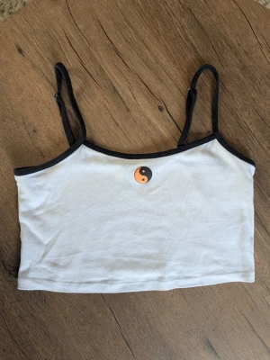 Vit linnetopp med yin yang-tryck - Säljer en vit linnetopp med svarta tunna axelband och svart kant. Toppen har ett litet orange och svart yin yang-tryck framtill. Perfekt till sommaren och enkel att matcha med jeans eller kjol.