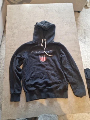 Mörkblå hoodie från GANT med logga - Snygg mörkblå hoodie från GANT med broderad logga och amerikanskinspirerat sköldmärke på bröstet. Tröjan har huva med vita dragsnören och en stor magficka. Tillverkad i mjukt bomullsmaterial som är skönt att bära.