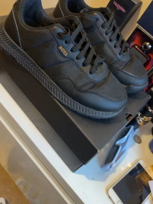 Svarta sneakers från BOSS - Stilrena svarta sneakers från BOSS med diskret logga på sidan. Skorna har en låg profil, snörning och en robust sula. Materialet är en mix av textil och syntet som ger en modern look. Perfekta för dig som gillar minimalistisk och clean stil.