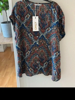 Mönstrad blus från ONLY - Säljer en färgstark blus från ONLY med ett detaljerat paisleymönster i blått, vinrött och beige. Blusen har rund halsringning och korta ärmar. Materialet känns lätt och luftigt, perfekt för en trendig vardagslook.