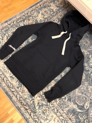 Svart hoodie från Random Bastards XS - Svart hoodie från Random Bastards med vit dragsko i huvan och stor magficka framtill. Tryck på ena ärmen och klassisk relaxed fit. Perfekt för dig som gillar streetwear och vill ha något bekvämt och stilrent.