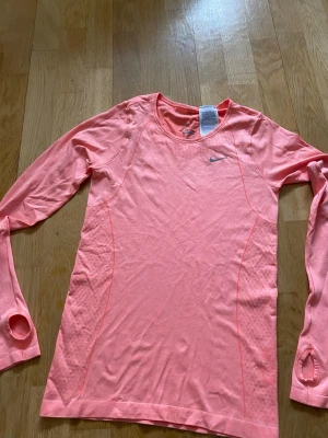 Nike rosa långärmad träningströja - Snygg långärmad träningströja från Nike i en fräsch rosa färg. Tillverkad i Dri-FIT-material som transporterar bort svett. Tröjan har tumhål vid ärmsluten och en tight passform som passar perfekt till löpning eller gym. Helt ny utan prislapp                                                    