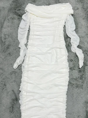 Vit långklänning med offshoulder - Elegant vit långklänning med offshoulder-design och långa ärmar. Klänningen har rynkade detaljer längs hela plagget som ger en snygg siluett. Tillverkad i ett mjukt och stretchigt material som sitter bekvämt på kroppen. Perfekt för speciella tillfällen.