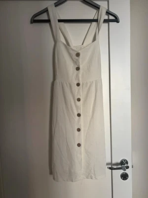 Vit sommarklänning med knappar - Säljer en vit klänning med breda axelband och knappar framtill. Klänningen har smock i ryggen för skön passform och är tillverkad i ett mjukt bomullstyg. Perfekt för varma dagar och enkel att styla med sneakers eller sandaler.