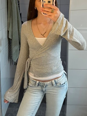 Grå omlottopp med knytband - Snygg grå omlottopp med långa ärmar och knytband i sidan. Tunn och mjuk kvalitet, perfekt för lager på lager. Djup V-ringning och figurnära passform ger en trendig look. Passar till både jeans och kjol för en avslappnad stil.