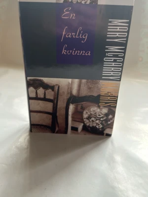 En farlig kvinna av Mary McGarry Morris - Romanen 'En farlig kvinna' av Mary McGarry Morris har ett omslag i blått och beige med bilder på en stol och blommor. Boken är inbunden och texten på omslaget är i vitt och lila. Perfekt för dig som gillar spännande skönlitteratur.