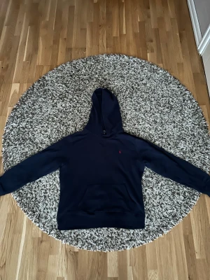 Mörkblå hoodie från Polo Ralph Lauren - Snygg mörkblå hoodie från Polo Ralph Lauren med klassisk huva och magficka. Diskret röd logga broderad på bröstet. Tillverkad i mjukt bomullsmaterial som känns skönt mot huden. Perfekt för en avslappnad och stilren look.