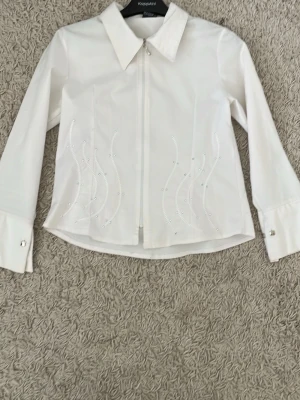 Vit skjorta med paljettdetaljer KappAhl - Stilren vit skjorta från KappAhl med lång ärm, klassisk krage och dragkedja framtill. Skjortan har snygga paljettdetaljer i vågigt mönster på framsidan och dekorativa knappar vid manschetterna. Perfekt för dig som gillar en clean look med lite extra bling.